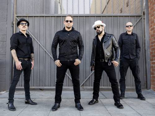 Die vier männlichen Bandmitglieder von Volbeat stehen Sonnenbrille-tragend vor einem Tor.